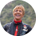 A woman smiling wearing a jacket and a leather motorcycle vest with patches over top of it.